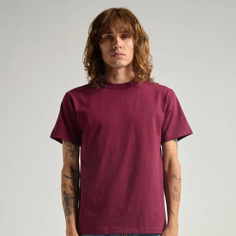 A man with long light brown hair and tattoos wearing a solid maroon crewneck t-shirt and blue jeans, standing against a light gray studio background.